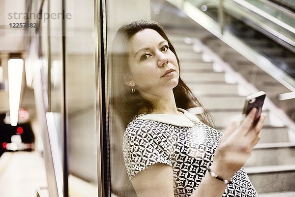 Junge Frau posiert mit Smartphone hinter einer Glaswand in einer U-Bahn-Station  Köln  Nordrhein-Westfalen  Deutschland  Europa