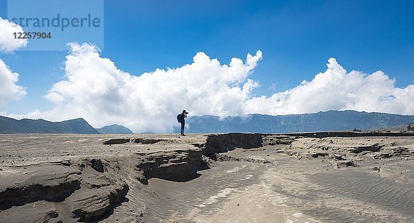 Junger Mann steht an einem Erdsplitter und fotografiert  rauchenden Vulkan Gunung Bromo  Mt. Batok  Mt. Kursi  Mt. Gunung Semeru  Tengger Caldera  National Park Bromo-Tengger-Semeru  Java  Indonesien.