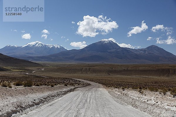 Schotterstraße führt durch das Altiplano  hinter schneebedeckten Vulkanen  bei Colchane  Tarapaca  Chile  Südamerika