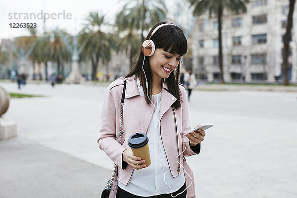 Spanien  Barcelona  lächelnde Frau mit Kaffee  Handy und Kopfhörer in der Stadt