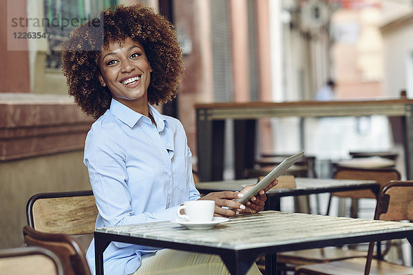 Lächelnde Frau mit Afro-Frisur sitzend im Outdoor-Café mit Tablette