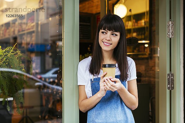 Lächelnde Frau steht vor der Eingangstür eines Ladens mit Kaffee zum Mitnehmen.