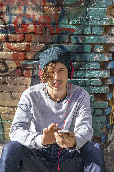 Porträt eines jungen Mannes mit Kopfhörer und Handy vor der Graffiti-Wand