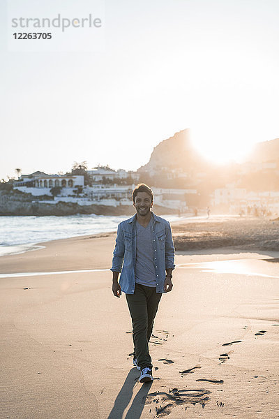 Glücklicher junger Mann  der bei Sonnenuntergang am Strand spazieren geht.