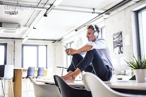 Geschäftsmann mit Smartphone und Kopfhörer  der barfuß auf dem Schreibtisch im Büro sitzt