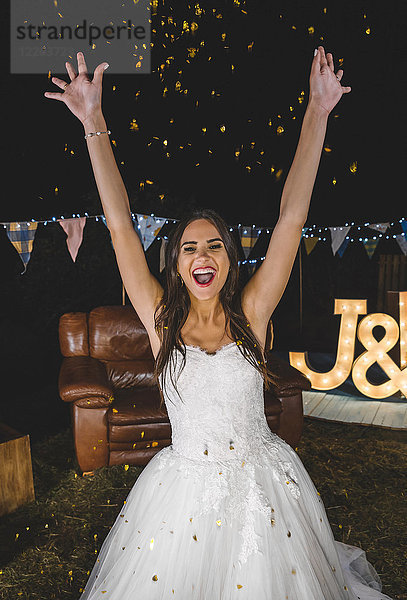 Fröhliche Braut  die ihre Arme hebt  während Konfetti auf einer Nachtparty im Freien über sie fällt.