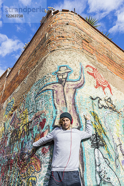 Junger Mann hört Musik mit Kopfhörern  die sich an die Graffiti-Wand lehnen.