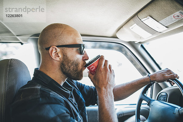 Junger Mann mit Sonnenbrille und Bart auf einem Roadtrip mit Takeaway-Getränk