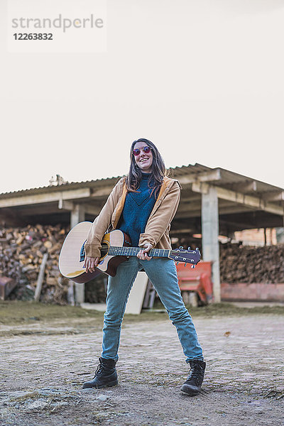 Lächelnde junge Frau hält Gitarre im Freien auf einem Bauernhof