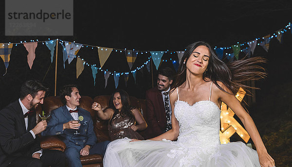 Fröhliches Brauttanzen auf einer nächtlichen Feldparty mit Bräutigam und Freunden im Hintergrund