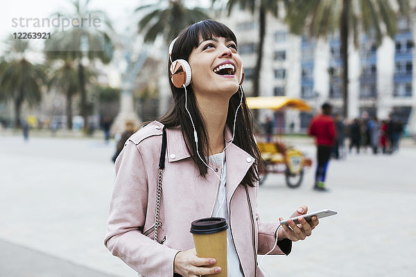 Spanien  Barcelona  lachende Frau mit Kaffee  Handy und Kopfhörer in der Stadt