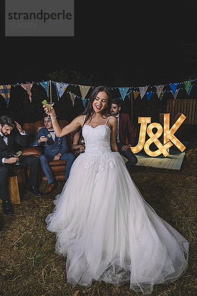 Fröhliche Braut mit Cocktailglas auf einer Nachtfeld-Party mit Bräutigam und Freunden im Hintergrund