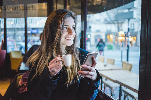 Paris  Frankreich  Porträt einer lächelnden jungen Frau mit Smartphone beim Espresso-Trinken in einem Café