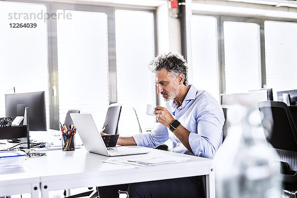 Erwachsener Geschäftsmann am Schreibtisch im Büro mit Laptop und Kaffeetrinken