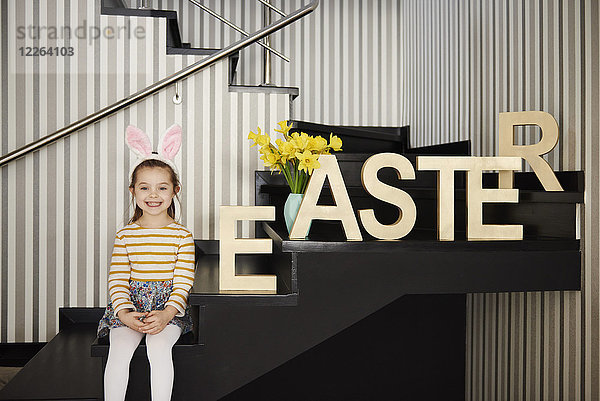 Porträt eines lächelnden Mädchens mit Hasenohren auf einer Treppe neben dem Wort'Ostern'.
