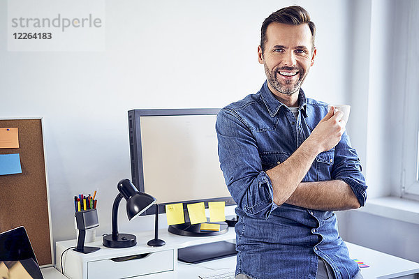 Porträt eines lächelnden Mannes mit Tasse Kaffee am Schreibtisch im Büro