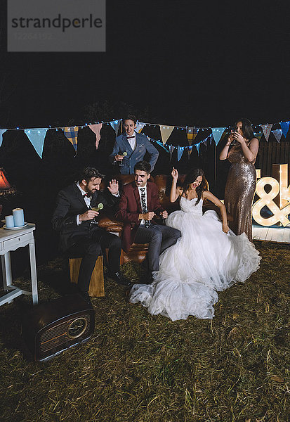 Glückliches Hochzeitspaar beim Sitzen auf dem Sofa mit seinen Freunden auf einer Nachtfeld-Party