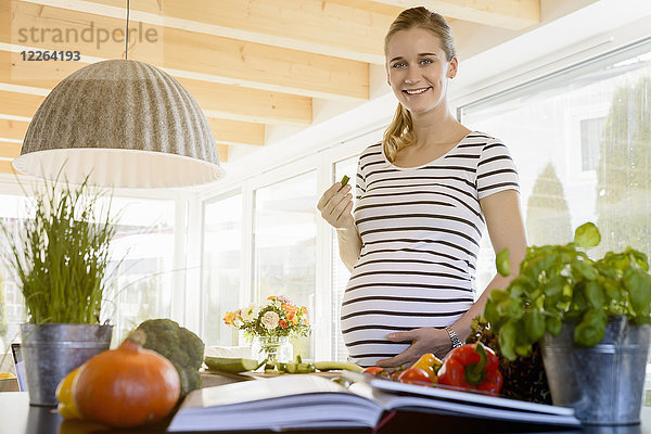 Porträt einer lächelnden schwangeren Frau in der Küche zu Hause beim Gurkenessen