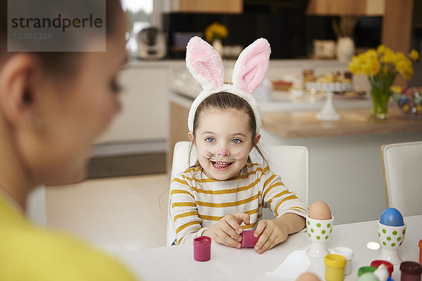 Mädchen mit Hasenohren und Mutter am Tisch sitzend mit Ostereiern