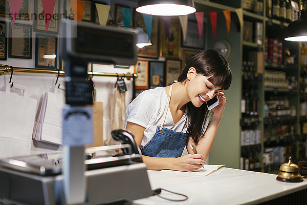 Lächelnde Frau am Telefon in einem Laden  die sich Notizen macht.