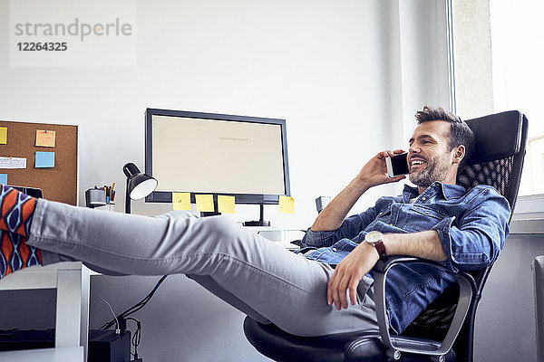 Entspannter Mann sitzt am Schreibtisch im Büro und telefoniert mit dem Handy.