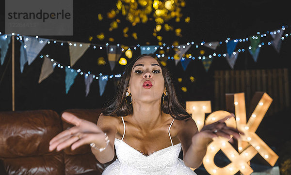Porträt einer glücklichen Braut  die Konfetti auf einer Nachtparty im Freien bläst.