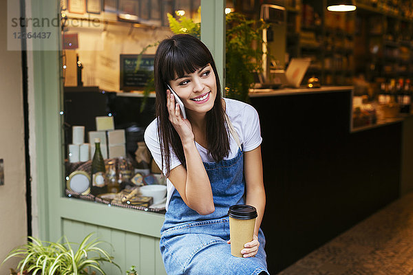 Lächelnde Frau sitzt an der Eingangstür eines Ladens und redet mit dem Handy.
