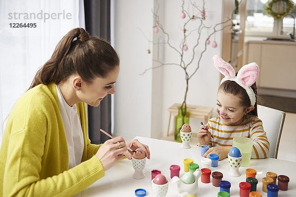 Glückliche Mutter und Tochter mit Hasenohren sitzen am Tisch und malen Ostereier.