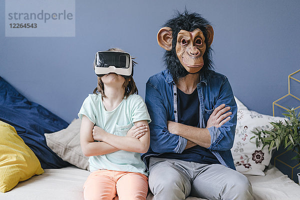 Vater mit Affenmaske sitzend neben Sohn mit VR-Brille zu Hause