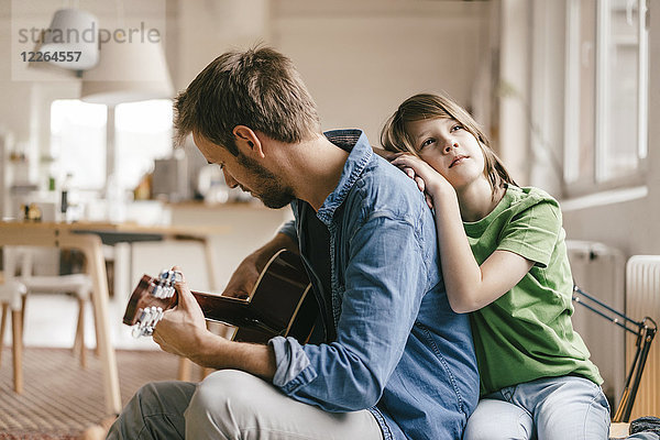 Sohn lehnt sich an Vater  der zu Hause Gitarre spielt.