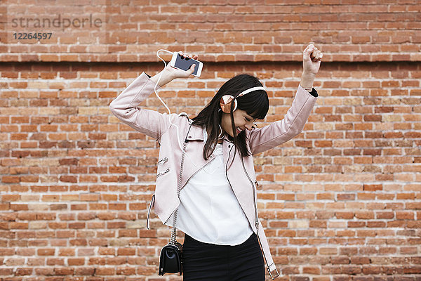 Fröhliche Frau mit Handy beim Musikhören über Kopfhörer an der Ziegelwand