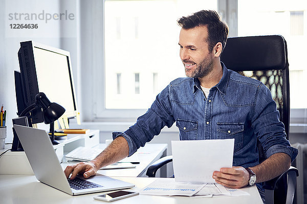 Lächelnder Mann mit Dokument und Laptop am Schreibtisch im Büro