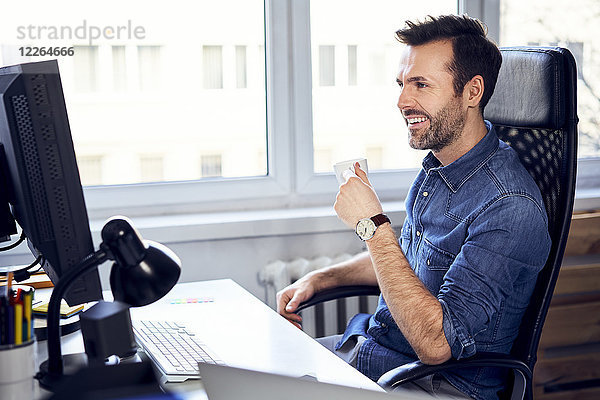 Lächelnder Mann schaut auf den Computer und trinkt Kaffee am Schreibtisch im Büro.