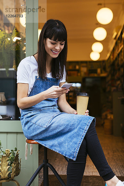 Lächelnde Frau auf Hocker sitzend mit Handy an der Eingangstür eines Ladens