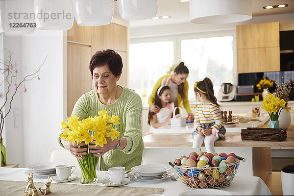 Seniorin arrangiert Blumen auf dem Esstisch mit Familie im Hintergrund