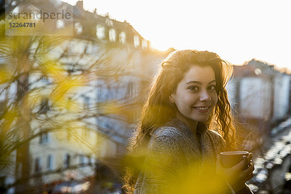 Porträt des lachenden Teenagermädchens mit Kaffeetasse auf dem Balkon bei Dämmerung