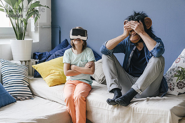 Vater mit Affenmaske sitzend neben Sohn mit VR-Brille zu Hause