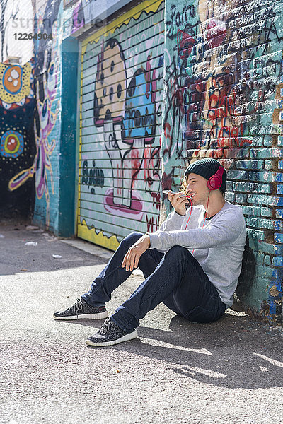 Junger Mann mit Kopfhörer vor der Graffiti-Wand sitzend mit Handy