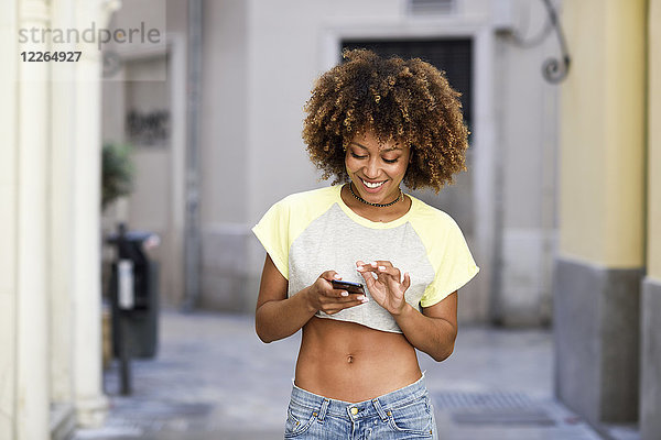 Lächelnde Frau mit Afro-Frisur mit Smartphone im Freien