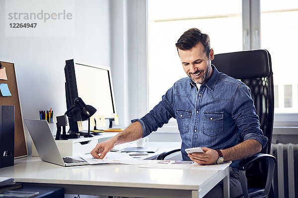 Lächelnder Mann mit Smartphone und Entwurf arbeitet am Schreibtisch im Büro