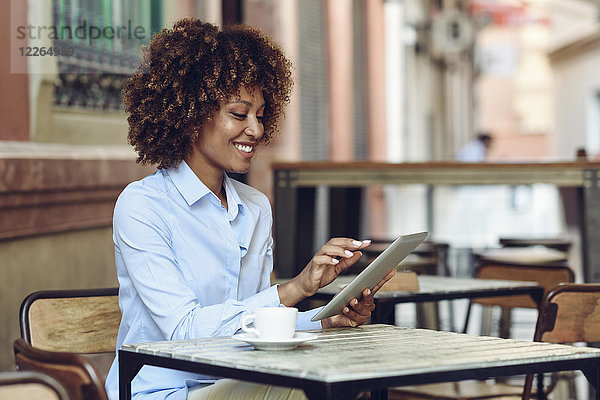 Lächelnde Frau mit Afro-Frisur sitzend im Outdoor-Café mit Tablette