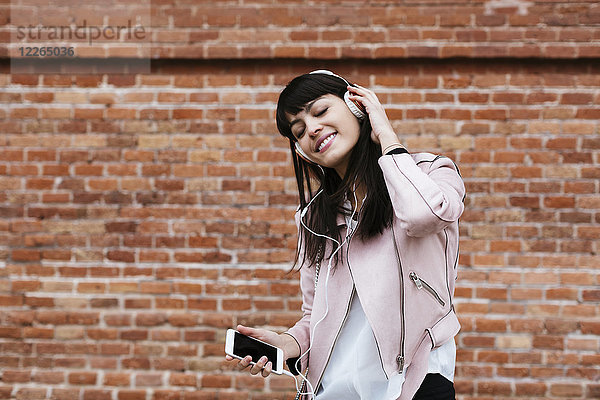 Lächelnde Frau mit Handy beim Musikhören über Kopfhörer an der Ziegelwand