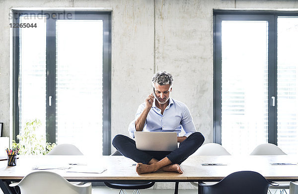 Reife Geschäftsleute sitzen barfuß auf dem Schreibtisch im Büro mit Handy und Laptop