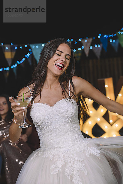 Fröhliche Braut lacht und tanzt  während sie einen Cocktail auf einer Party im Freien hält.
