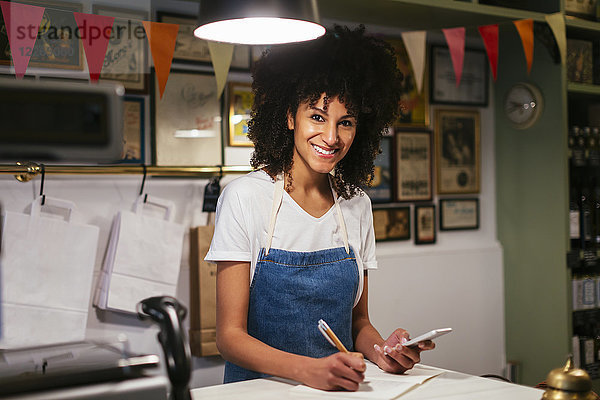 Porträt einer lächelnden Frau mit Handy und Notizbuch im Laden