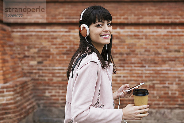 Porträt der lächelnden Frau mit Kaffee  Handy und Kopfhörer an der Ziegelwand