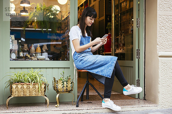 Frau auf Hocker sitzend mit Handy an der Eingangstür eines Ladens