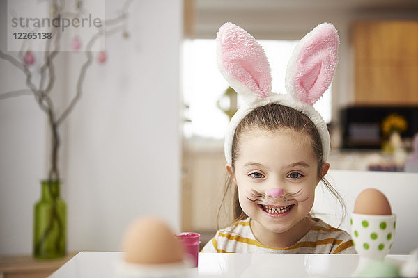 Porträt eines lächelnden Mädchens mit Hasenohren am Tisch mit Ostereiern