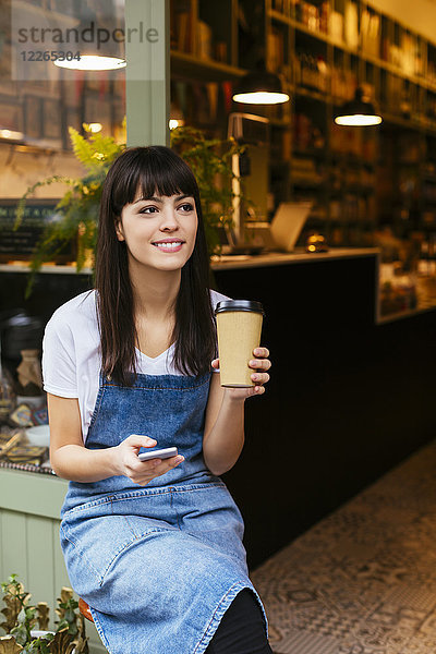 Lächelnde Frau sitzt an der Eingangstür eines Ladens mit Handy und Kaffee zum Mitnehmen.