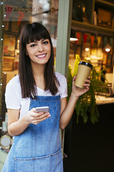Porträt einer lächelnden Frau mit Handy und Kaffee zum Mitnehmen in der Eingangstür eines Ladens